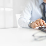 A suited professional seated at a desk, using a calculator to work on financial figures, with a pair of glasses and documents nearby.