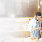 A smiling man and woman wearing striped aprons stand behind a bakery counter, looking down together at a tablet. Baked goods are displayed in front of them, and the café-style background is softly lit and welcoming.
