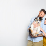 A smiling man and woman stand close together against a plain white background. The man, dressed in a light blue shirt and khaki pants, holds an adorable fluffy puppy in his arms. The woman, wearing a mustard-yellow sleeveless top, reaches over to gently pet the pup while looking at it with affection. The puppy is a tricolor—mostly white with black and tan markings—and gazes forward.