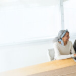 Smiling mature couple sitting at a wooden table in a bright, airy room; the woman works on a laptop while the man, holding a coffee mug, looks on warmly.