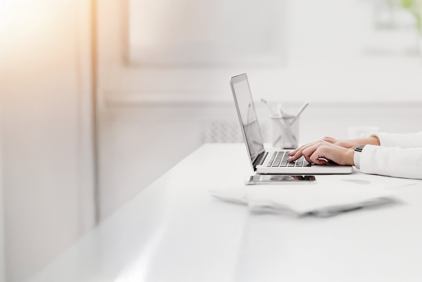 Person typing on a laptop at a clean white desk with sunlight streaming in, minimal office setup with papers, a phone, and a pen holder nearby.