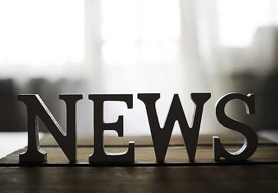 Photo of block letters spelling NEWS in white or light-colored material, standing upright on a dark table or wooden surface. The background is softly blurred with light coming through windows, giving a calm, slightly out-of-focus ambience.