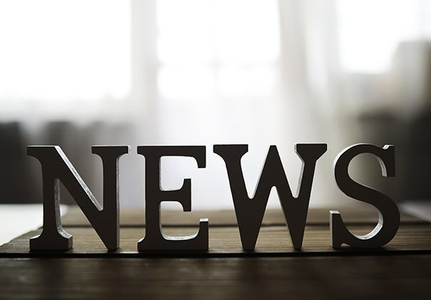 Photo of block letters spelling NEWS in white or light-colored material, standing upright on a dark table or wooden surface. The background is softly blurred with light coming through windows, giving a calm, slightly out-of-focus ambience.