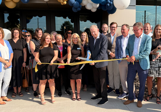 Group of men and women in business attire pose for a ribbon-cutting ceremony outside a new building, with a man in the center using large scissors to cut a blue and yellow ribbon. Blue, gold, and white balloon arches frame the entrance behind them.