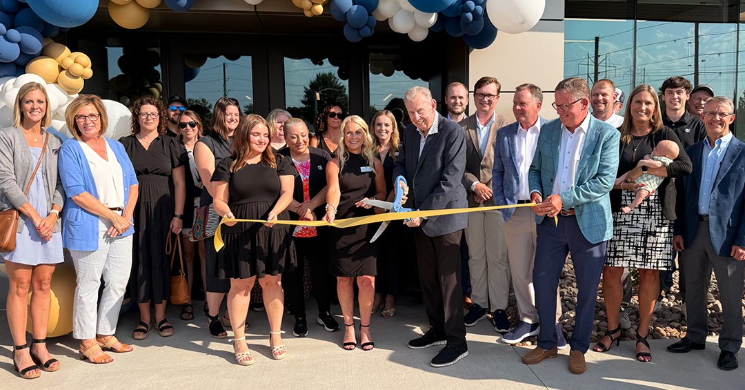 Group of men and women in business attire pose for a ribbon-cutting ceremony outside a new building, with a man in the center using large scissors to cut a blue and yellow ribbon. Blue, gold, and white balloon arches frame the entrance behind them.